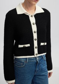 Schwarze strukturierte Strickjacke mit cremefarbenem Kragen, knöpfbarem Vorderteil und zwei Fronttaschen. Mit großen metallischen Knöpfen und kontrastierenden Saumdetails.