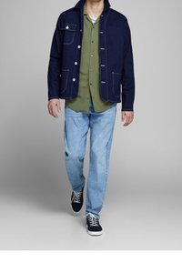 Navy denimjack met witte stiksels, gecombineerd met een groen shirt en lichtblauwe jeans. Schoeisel bestaat uit donkere sneakers met witte zolen.