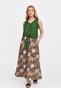 Zielony bezrękawnik z dekoltem w serek i detalami guzików, w zestawieniu z długą brązową spódnicą z białym kwiatowym wzorem. Czarny sandały.