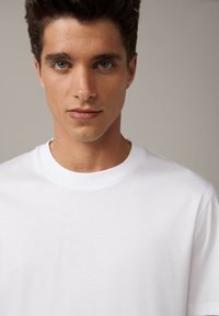 T-shirt bianco in cotone con colletto a girocollo, caratterizzato da una texture liscia e un fit classico, parzialmente visibile sul busto di un modello.