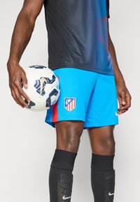 Blå fodboldshorts med røde detaljer og et Atlético Madrid-logo, parret med sorte knæhøje sokker, som holder en mønstret hvid fodbold.