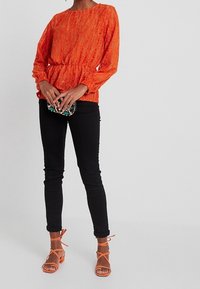 Orangefarbener Pullover mit Struktur und gerafftem Taillenbereich, kombiniert mit schwarzen Skinny-Jeans und orangefarbenen Riemchensandalen. Accessoire: kleine verzierten Clutch.