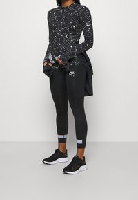 Schwarzes, sportliches Langarmoberteil mit geometrischem weißen Muster, kombiniert mit schwarzen Leggings mit subtilen Mustern und reflektierenden Akzenten sowie schwarzen Turnschuhen.