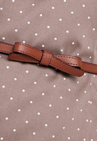 Tissu marron à pois blancs, avec une ceinture en simili cuir marron nouée en nœud à la taille. Texture lisse avec un éclat minimal.