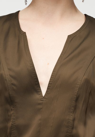 Λαδί πράσινη μπλούζα με βαθύ V λαιμόκοψη, με διακριτικές ραφές και απαλό, ελαφρύ ύφασμα που πέφτει απαλά.