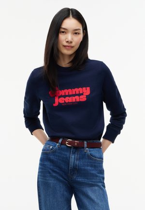 Jeune femme aux longs cheveux noirs portant un sweat bleu marine avec un texte rouge et un jean bleu, debout les mains derrière le dos.