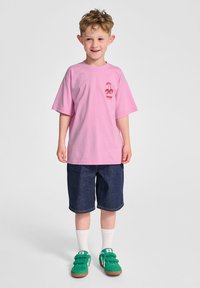 Rosa bomullströja med röd körsbärsgrafik, kombinerad med mörka denimshorts och gröna sneakers. Vita strumpor synliga ovanför skorna.