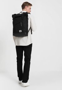 Mochila negra tipo roll-top fabricada con materiales de tela y plástico. Cuenta con correas ajustables, un bolsillo lateral con cremallera y un parche de logo blanco.
