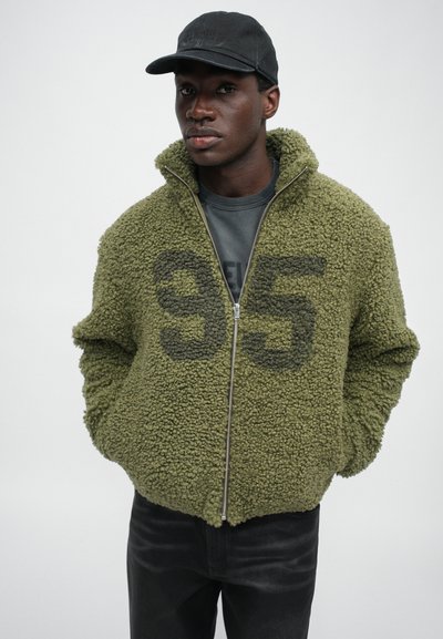 Olive green, zip-up fleece jacket with a textured, shaggy finish and a large gray “95” printed on the front. Black cap, dark jeans.