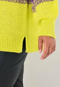 Maglione giallo lavorato a maglia con orlo a coste, caratterizzato da accenti a motivo viola. Dettagli con spacco laterale visibili, abbinato a pantaloni di pelle nera.