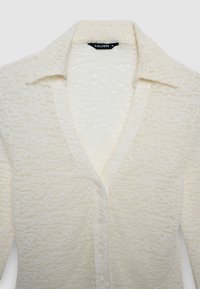 Strukturierte, cremefarbene Bluse mit Kragen, Knopfleiste und dezentem Muster. Der Stoff wirkt leicht und leicht zerknittert.