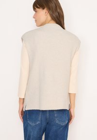 Beige gerippter Sweater-Weste mit hohem Kragen, Schulternähten und seitlichen Schlitzen. Über einem hellen Langarmoberteil getragen, kombiniert mit blauen Jeans.