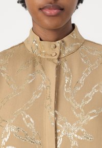Blusa beige con colletto alto, due bottoni e un motivo a catena metallica dorata. Il tessuto è liscio con una leggera lucentezza. Nessun dettaglio di chiusura visibile.