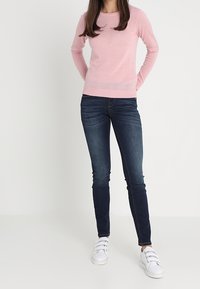 Un pull long à manches longues rose clair, avec une texture côtelée, associé à un jean skinny bleu foncé et des baskets blanches avec trois sangles réglables.