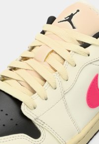 Κοντινή λήψη ενός αθλητικού παπουτσιού με λευκά κορδόνια, μαύρη μύτη, μπεζ γλώσσα, ροζ λογότυπο Nike και μαύρο λογότυπο Jumpman στη γλώσσα.