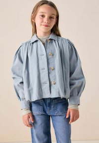 Jeune fille avec de longs cheveux portant une veste bleu clair boutonnée, manches de chemise rayées visibles, et jeans bleus, se tenant devant un fond beige.