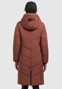 Person iført en lang brun quilted frakke med hætte og ryglynlås, parret med en sort kasket og sorte bukser, vendt væk fra kameraet.