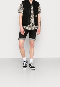 Gilet senza maniche nero su una camicia a maniche corte beige e nera con fantasia, abbinato a shorts in denim nero arrotolati e scarpe da ginnastica nere con accenti bianchi.