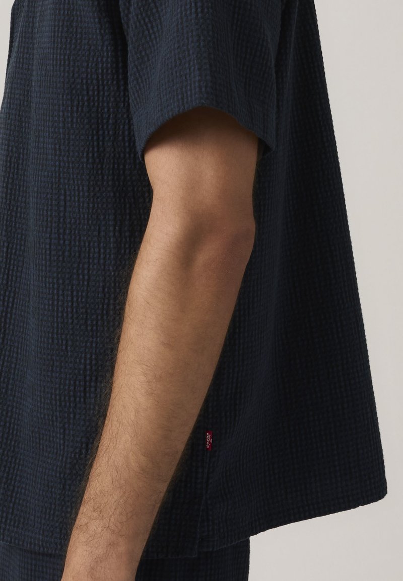 Mandearm synlig iført en kortærmet, struktureret mørkeblå skjorte med en lille rød stofmærkat nær kanten.