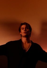 Jonge man in een zwarte open voorzijde shirt met een V-hals, staande tegen een warme oranje achtergrond, met uitgestrekte armen, en meerdere lagen kettingen.