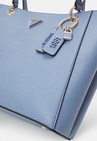 Jasnoniebieska torebka teksturowana z złotymi detalami, z plakietką logo oraz dwoma brelokami z napisami "GUESS" i "1981".