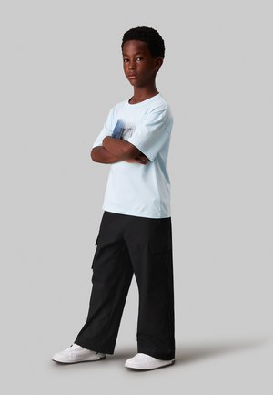 Maglietta azzurra chiara con un design grafico, abbinata a pantaloni cargo neri con tasche laterali e sneaker bianche.