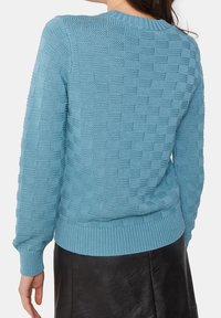 Pull turquoise en tricot avec un motif quadrillé texturé, col et poignets côtelés, associé à une jupe en cuir noire.