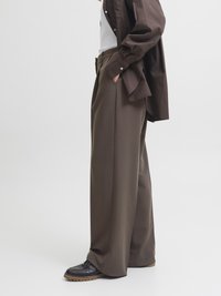 Calças largas, castanhas, feitas de tecido suave, com bolsos laterais. Combinadas com sapatos de couro pretos e uma camisa escura.