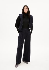 Marineblauwe gestreepte broek met hoge taille, gecombineerd met een zwarte coltrui en een bijpassend mouwloos vest dat over de schouders is gedrapeerd.