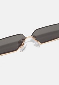 Sechseckige Sonnenbrille mit grauen Gläsern, goldfarbenem Metallrahmen und transparenten Nasenpads. Merkmale ein dünnes, geometrisches Design.