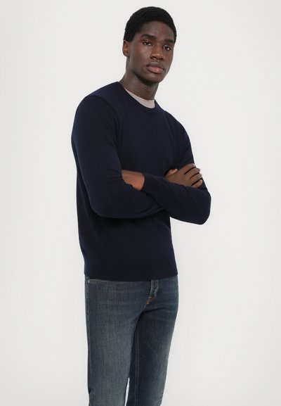 Maglione lavorato a maglia blu navy con scollatura a girocollo e polsini e orlo a costine. Indossato con jeans in denim blu, il modello ha le braccia incrociate.