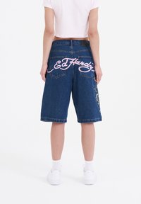 Shorts de mezclilla con un lavado oscuro, que presentan un logo bordado en rosa de "Ed Hardy" en la parte trasera y diseños intrincados en los laterales.