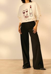 Sweatshirt crème avec des graphiques de vin rouge, associé à un pantalon noir ample et des chaussures noires faciles à enfiler, sur un fond neutre.