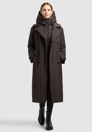 Femme portant un long manteau sombre à double boutonnage par-dessus une veste matelassée avec un col haut et des bottines noires, debout devant un fond uni.