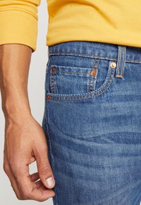 Vaqueros de mezclilla azul con un diseño clásico de cinco bolsillos, remaches de cobre y ligeras marcas de desgaste en la superficie, combinados con una camisa amarilla de manga larga.