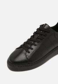Basket en cuir noir avec lacets noirs, finition lisse, détails cousus et semelle épaisse sur un fond clair uni.
