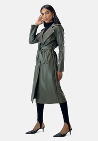 Zielono-oliwkowy płaszcz trench z skóry ekologicznej z paskiem w talii, kołnierzem typu lapel i kieszeniami bocznymi. Nosi się go na czarnym golfie i legginsach.