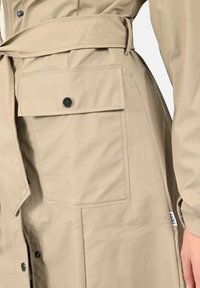 Μπεζ μακρύ παλτό από λεία, αδιάβροχη ύλη με ζωνάκι στη μέση, μεγάλη τσέπη με καπάκι και μαύρα κουμπώματα για κλείσιμο.