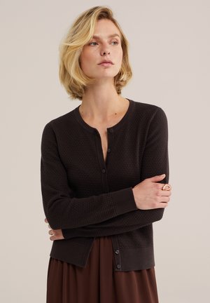 Blonde Frau trägt eine dunkel strukturierte Strickjacke über einem braunen Rock, Arme verschränkt, schaut zur Seite vor einem einfarbigen Hintergrund.