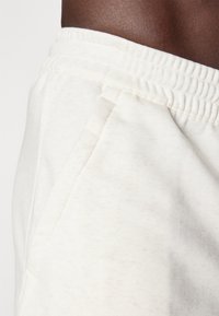 Cream-colored sweatpants feature a stretchy waistband and a side pocket with a soft texture and subtle speckled pattern.