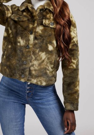 Braun-cremefarbene Jacke mit Fellmuster und Knöpfen sowie Klappentaschen, getragen über einem weißen Top, kombiniert mit hoch taillierten blauen Jeans mit sichtbaren Knöpfen.