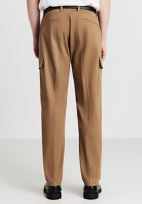 Pantaloni cargo color sabbia con vestibilità rilassata, dotati di tasche laterali, tasche posteriori piatte e una texture liscia. Abbinati a scarpe nere.
