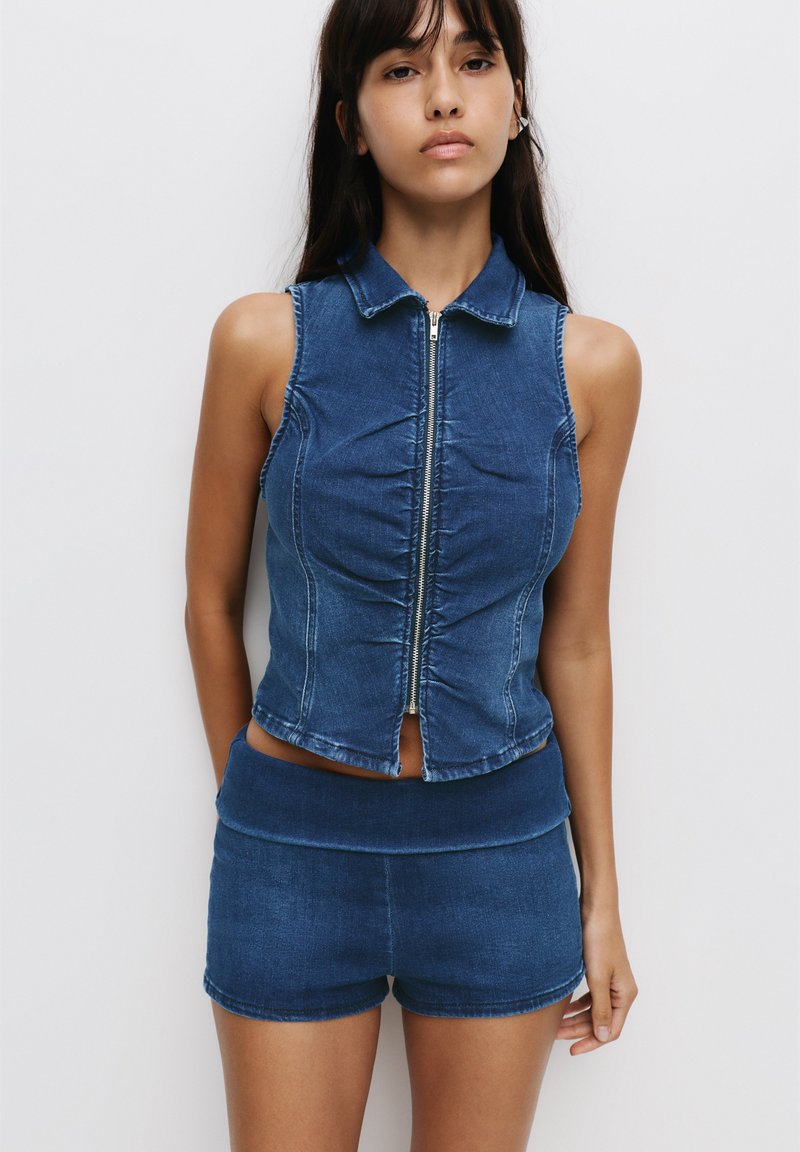 Denim mouwloze top met een kraag en een rits aan de voorkant, gecombineerd met bijpassende denim shorts met hoge taille. Beide in een middelblauwe wassing.