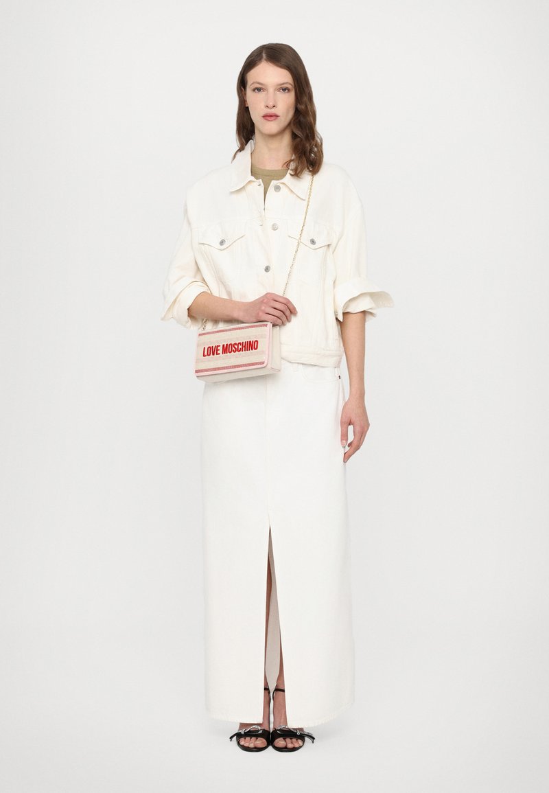 White denim jacket over a beige top, paired with a long white skirt featuring a front slit. Holding a light pink bag with red lettering.