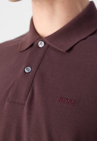 Gros plan d'une personne portant un polo brun rougeâtre foncé avec deux boutons et "BOSS" brodé sur la poitrine.