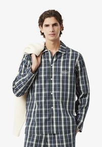 Set di pigiama in cotone a scacchi nei colori navy, verde e bianco. Camicia con bottoni, maniche lunghe e dettaglio con taschino. Il modello tiene in mano un maglione in maglia.