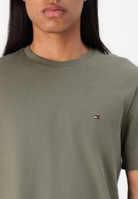 T-shirt di cotone verde oliva con colletto rotondo, maniche corte e un piccolo logo ricamato in rosso, bianco e blu sul lato sinistro del petto.