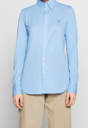 Femme portant une chemise bleu clair à manches longues boutonnée avec col et un pantalon beige, debout devant un fond uni.