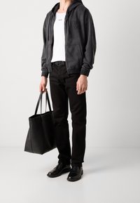 Schwarze Kapuzen-Zip-Up-Sweatjacke mit Logo, weißes T-Shirt mit Rundhalsausschnitt, schwarze Hose und glänzende schwarze Slipper. Trägt eine große schwarze Tote-Tasche.