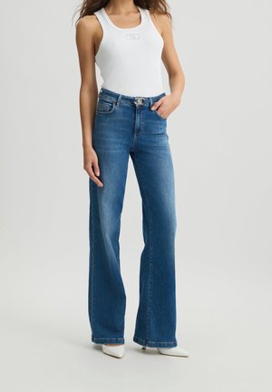 Femme portant un haut blanc sans manches et un jean bleu taille haute à jambes larges, avec des talons blancs pointus, debout avec une main dans la poche.